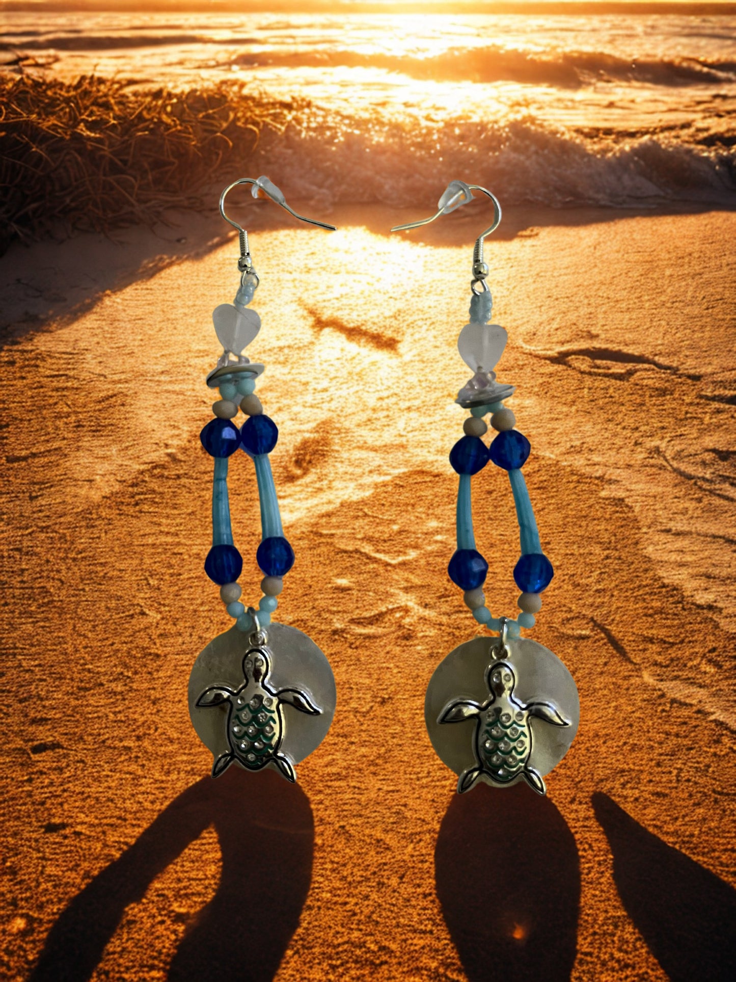 Turtle-themed earrings with blue and green beads on a sandy beach at sunset.