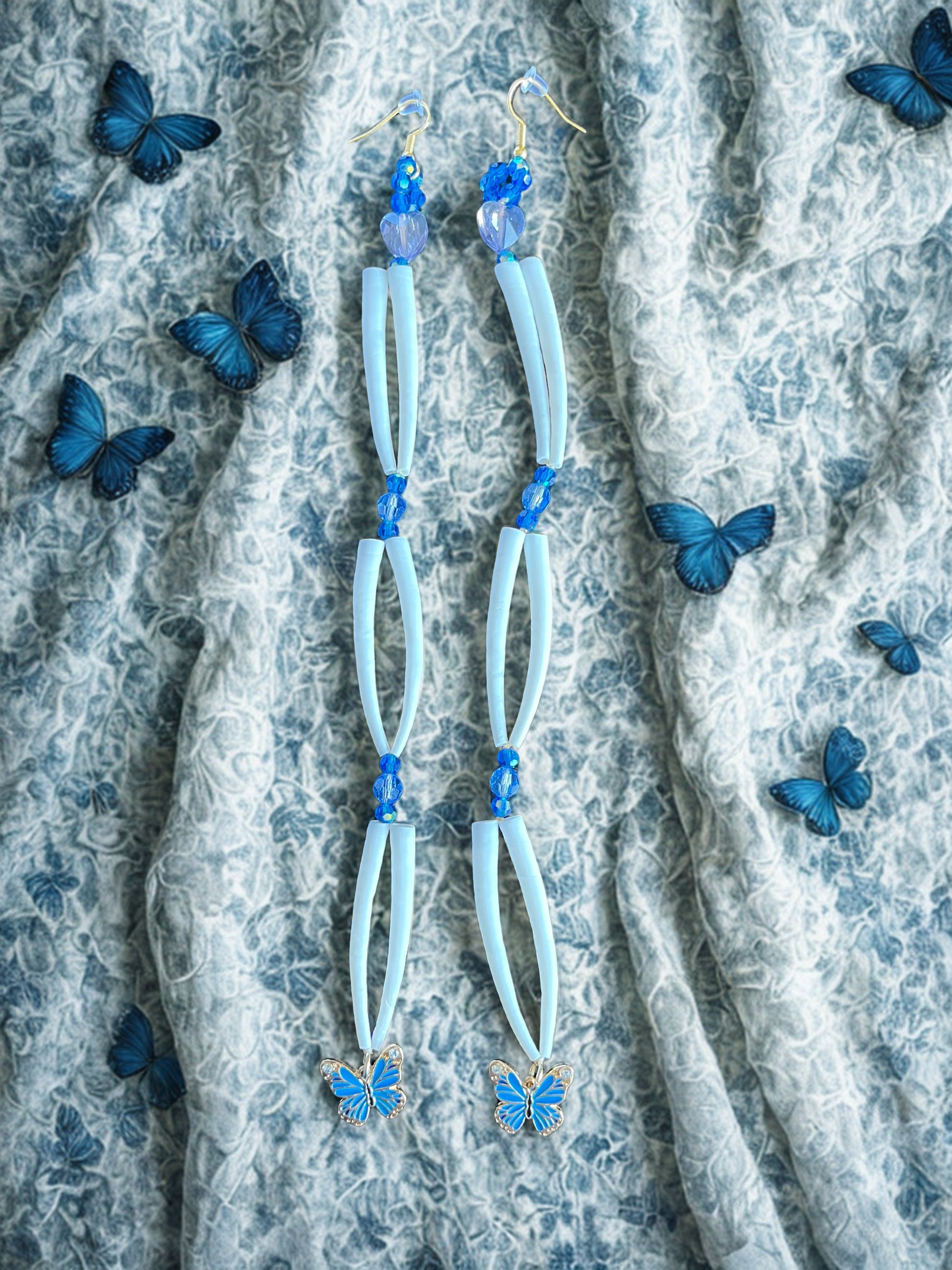 Light blue earrings with blue butterfly charms on a textured fabric background with additional blue butterflies.