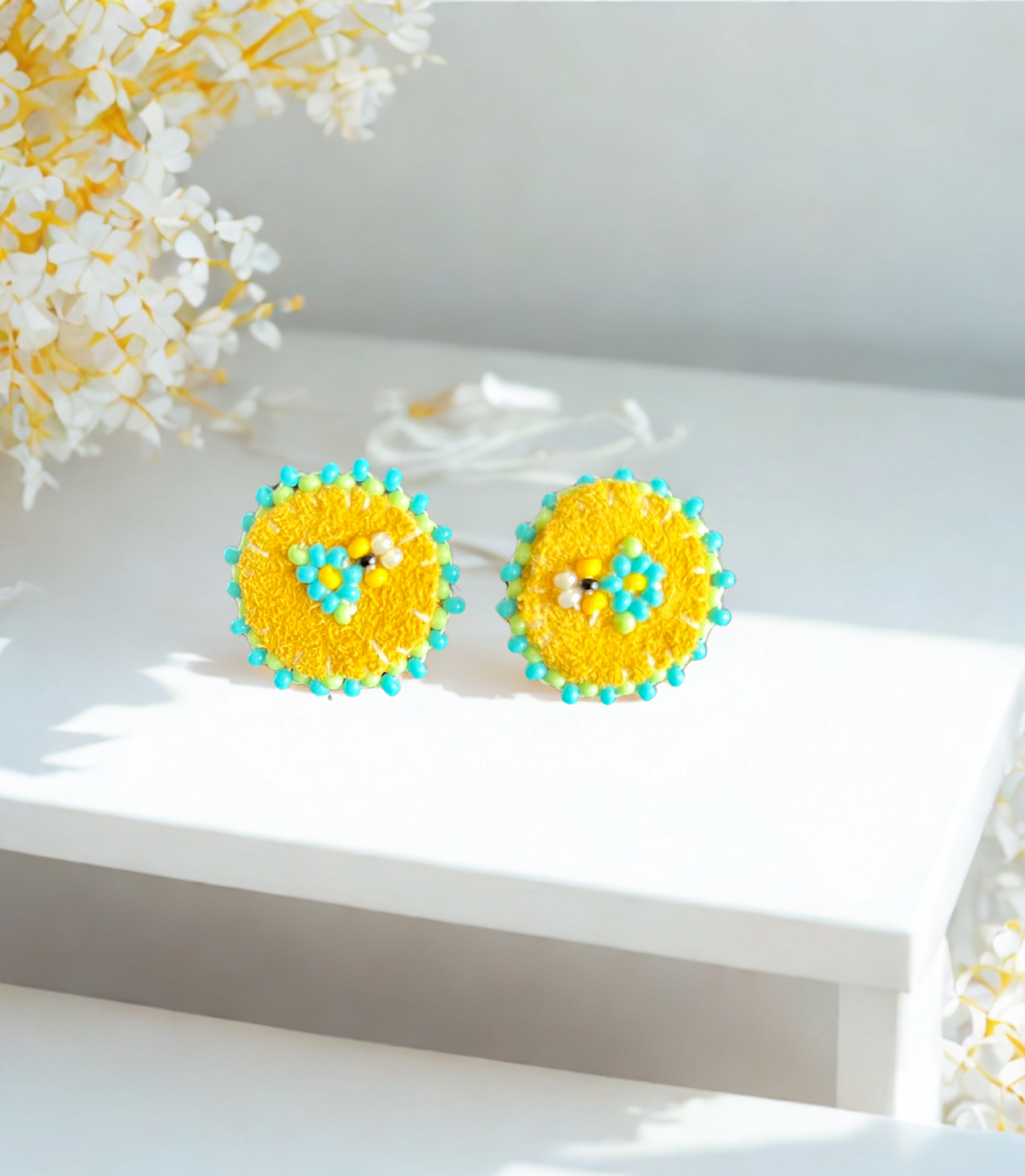 Yellow and blue circular earrings on a white surface with flowers in the background