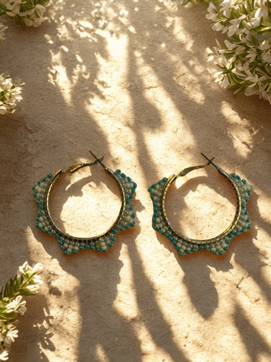 Pair of gold hoop earrings with green beaded accents on a sandy surface with white flowers.