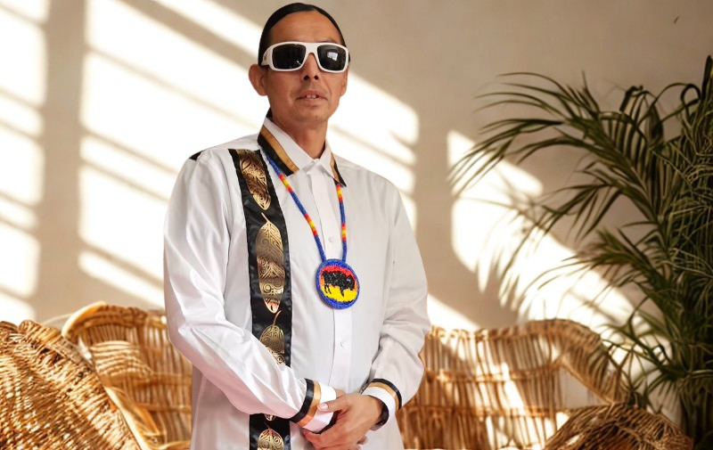 Person wearing sunglasses and a white shirt with decorative elements, standing indoors with plants in the background.