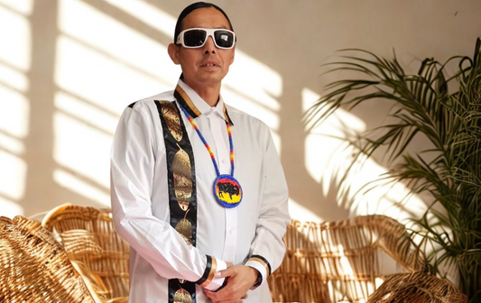 Person wearing sunglasses and a white shirt with decorative elements, standing indoors with plants in the background.